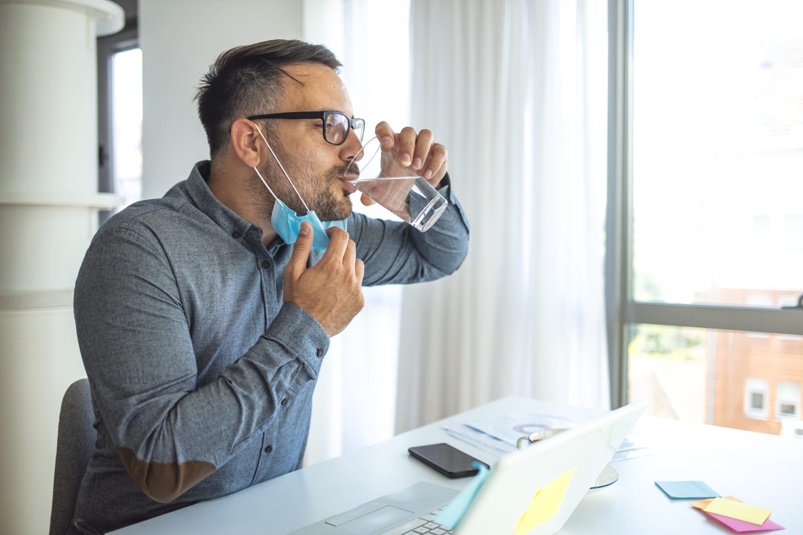 Businessman with mask drinking water at work. Staying hydrated on his business. Businessman having a glass of water while working in office and looking away - dehydration, drink enough liquids throughout the day